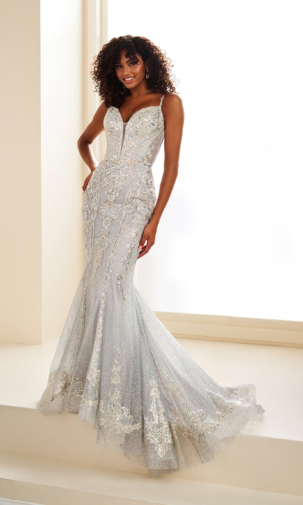 A woman in the Ellie Wilde EW36056 long prom dress, a fitted silver mermaid gown with lace applique and sequins, stands on a light staircase before a bright window.